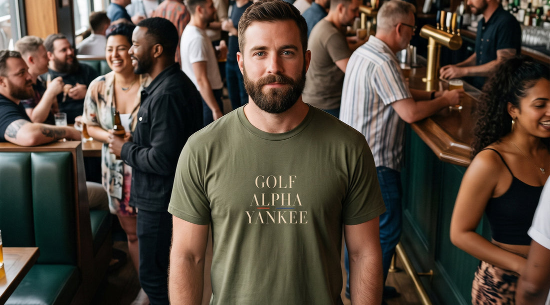 Military-green Golf Alpha Yankee Pride T-shirt with cream serif lettering and a subtle rainbow accent spelling G-A-Y in NATO code.