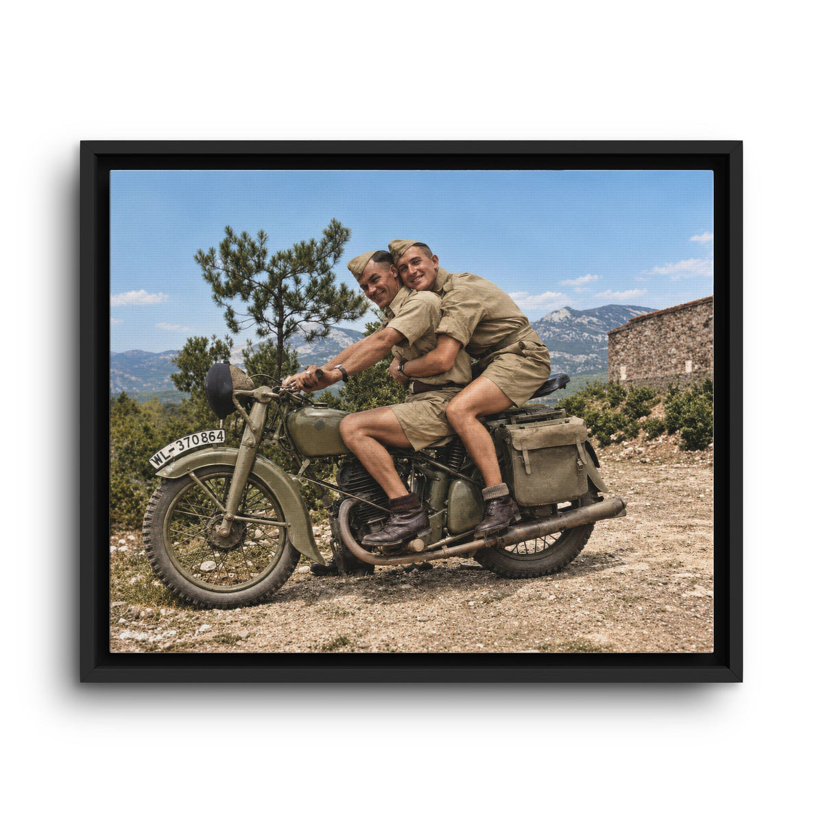 World War II-era motorcycle portrait showing two men in warm-weather uniforms with a stone building and mountains behind them.