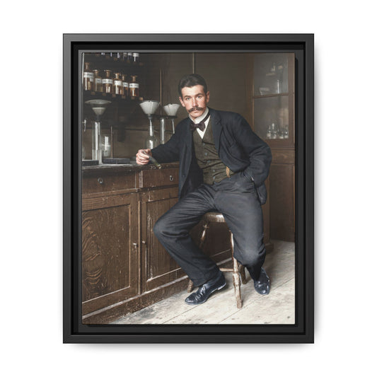 Colorized historical photograph of a moustached man in a dark suit and bow tie, seated on a stool holding a glass tube among laboratory glassware and wooden cabinets.