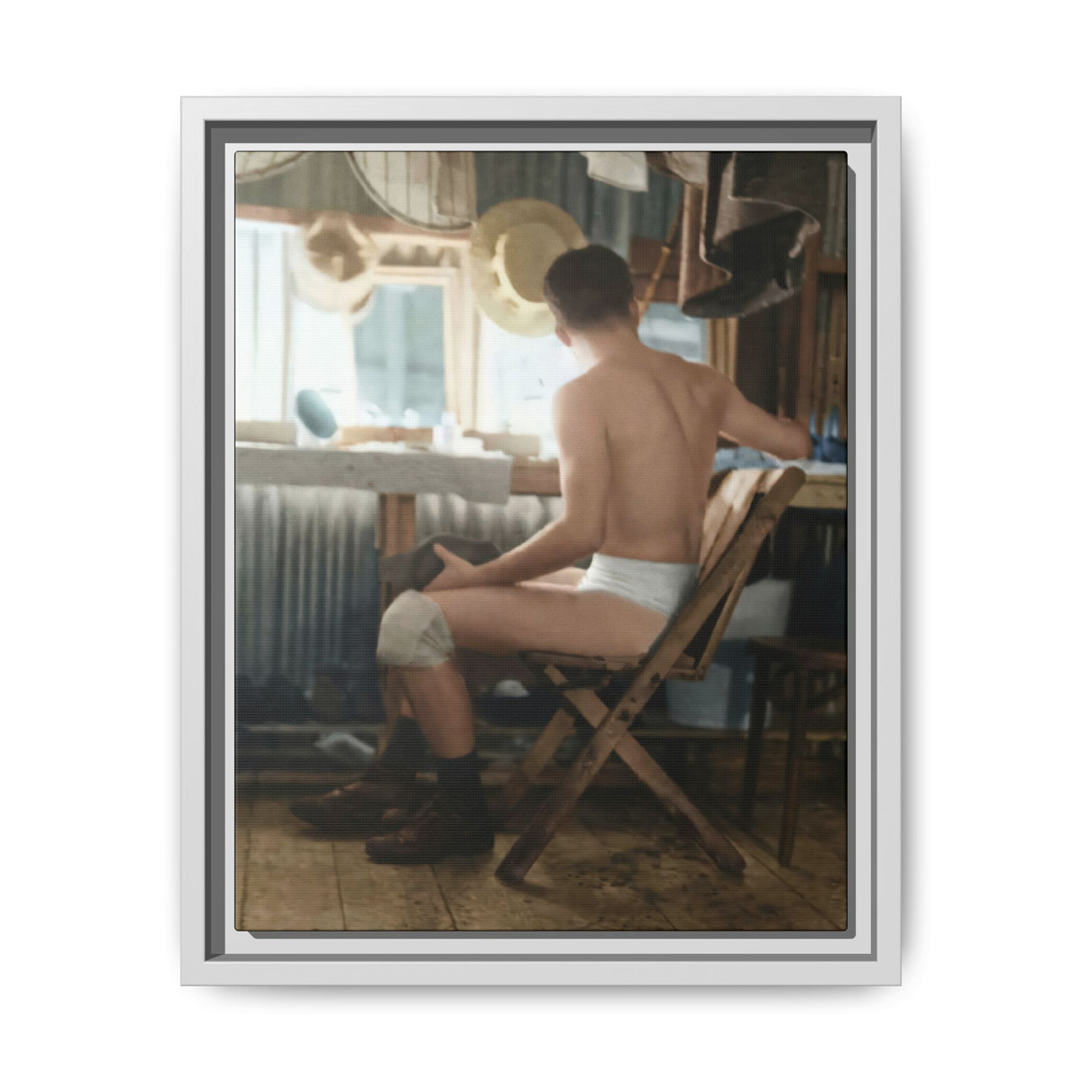 Restored colorized photograph of a bare-chested man viewed from behind on a wooden folding chair, with a mirror, hanging hats, radiator, and worn floorboards.