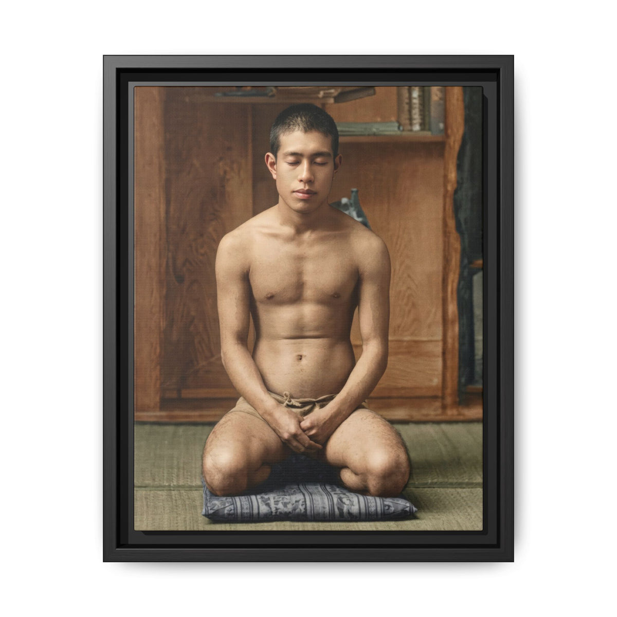 Colorized portrait of a bare-chested man kneeling on a patterned cushion, hands together in his lap and eyes appearing closed, before wooden shelves.