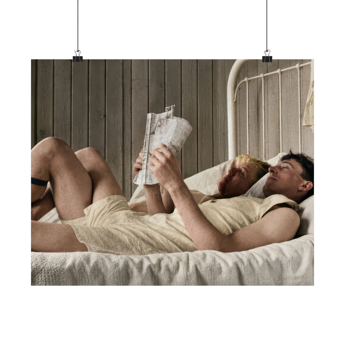 Two men recline together on a bed in a wood-paneled room; one holds a newspaper while the other rests against a pillow behind him.