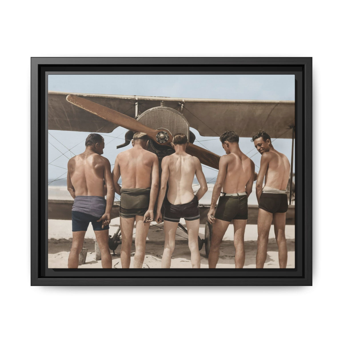 Restored colorized photograph of five bare-chested men in short trunks, seen from behind before a biplane on sandy ground.