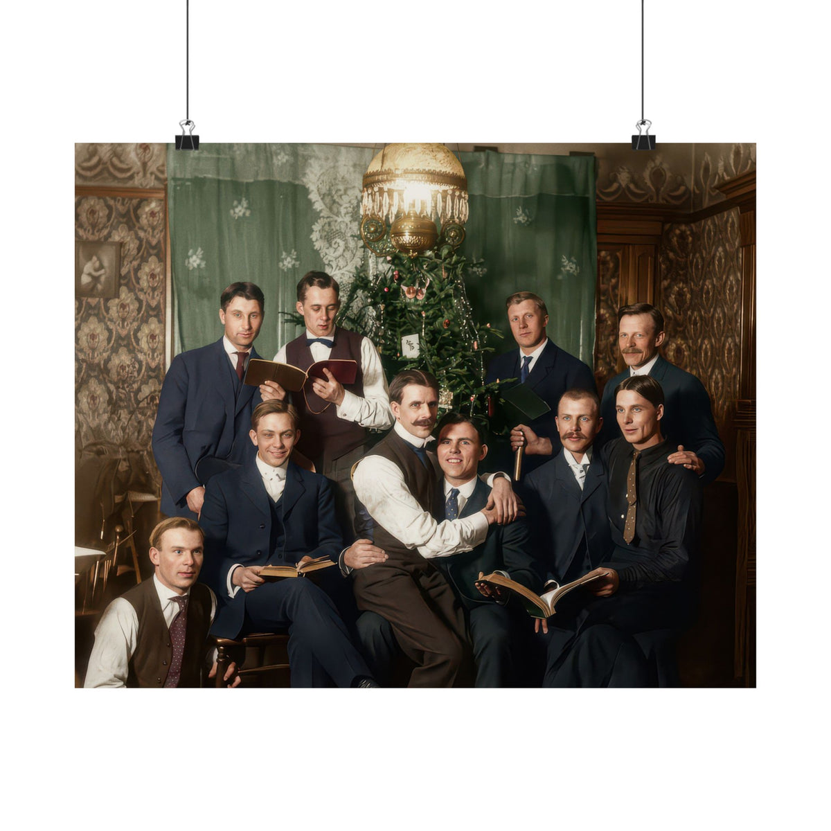 Nine men in early twentieth-century suits and waistcoats gather around a decorated Christmas tree, several holding open books.