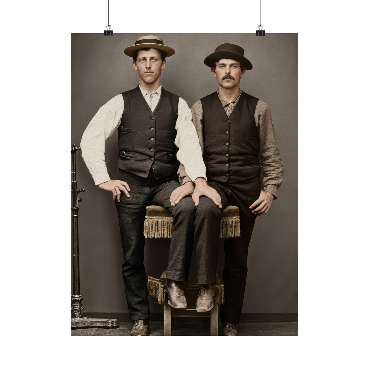 Two Victorian men in waistcoats and brimmed hats posed around a fringed studio stool, circa 1890–1900.